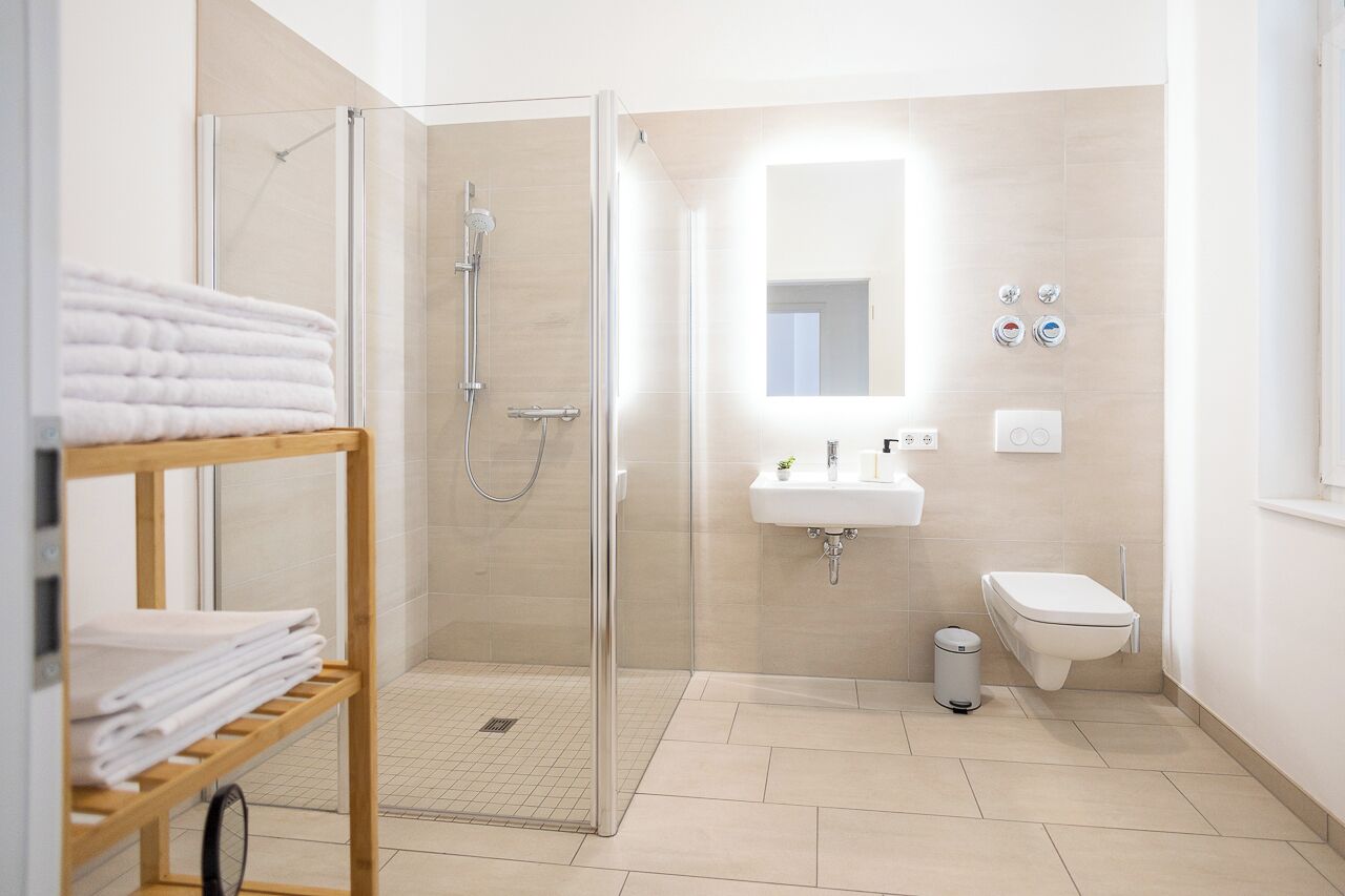 Ein modernes Badezimmer mit einer gläsernen Duschkabine, einem Waschbecken und einer Toilette. Ein Holzregal bietet gefaltete Handtücher und einen Mülleimer.