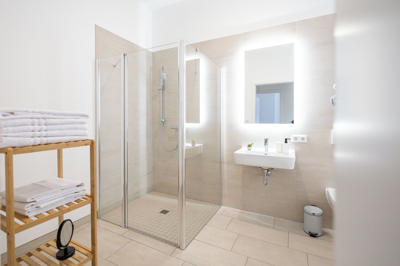 Une salle de bain moderne avec une douche à parois de verre, une étagère en bois où sont posées des serviettes, un lavabo blanc et une petite poubelle.
