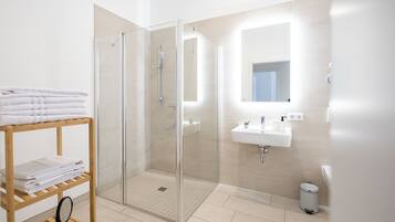Une salle de bain moderne avec une douche à parois de verre, une étagère en bois où sont posées des serviettes, un lavabo blanc et une petite poubelle.