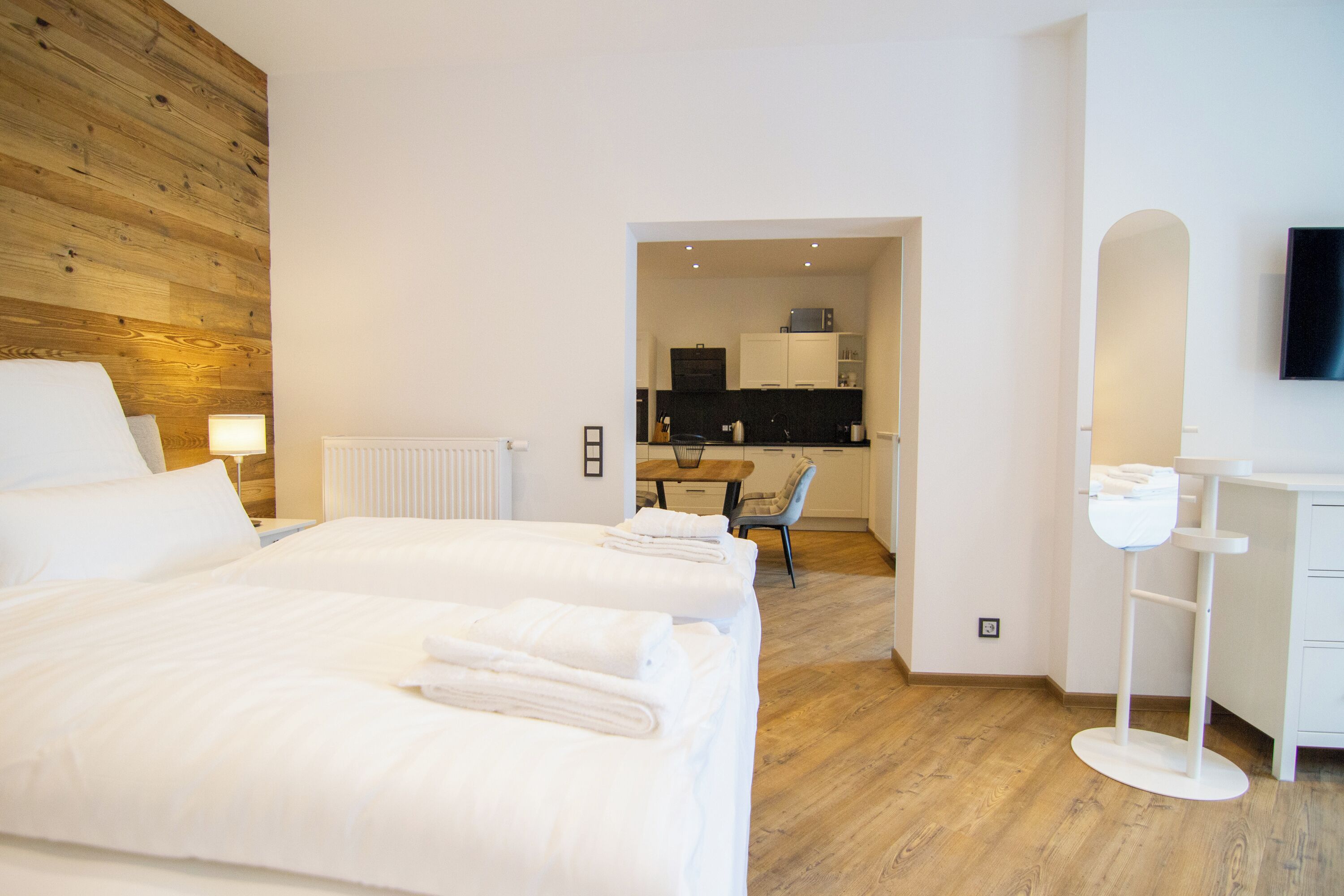 Quarto de hotel com duas camas, parede de destaque em madeira, cozinha compacta e televisão fixada na parede.