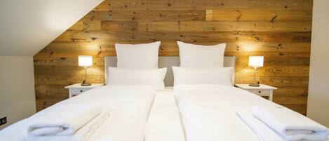 A double bed with white linens and towels, two bedside tables with lamps, and a wooden sloped ceiling.
