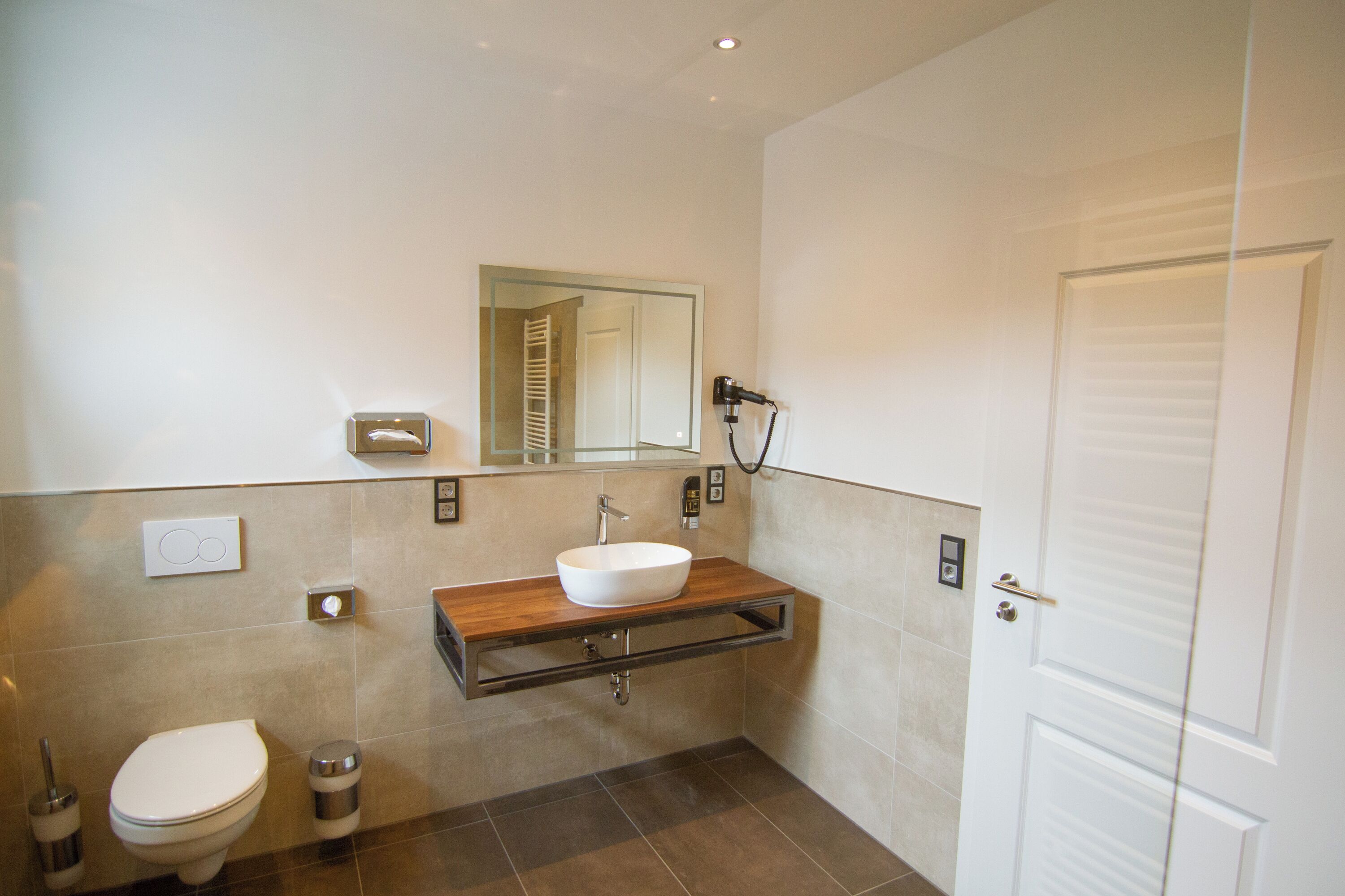 Un baño moderno con inodoro blanco, tocador de madera con lavamanos blanco y espejo sobre el lavamanos.