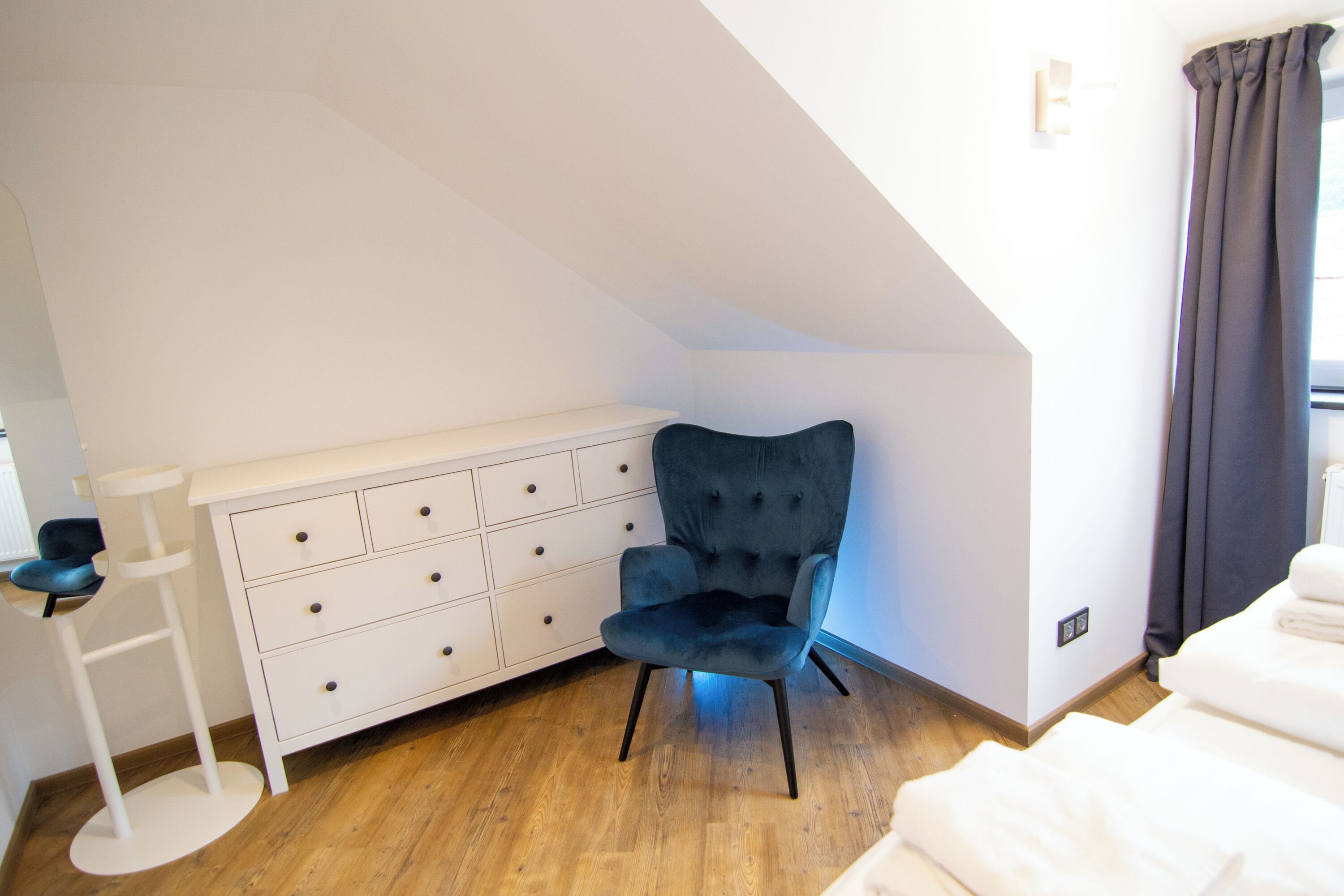 Un dormitorio con techo inclinado, un sillón tapizado azul oscuro, un tocador blanco y suelo de madera.