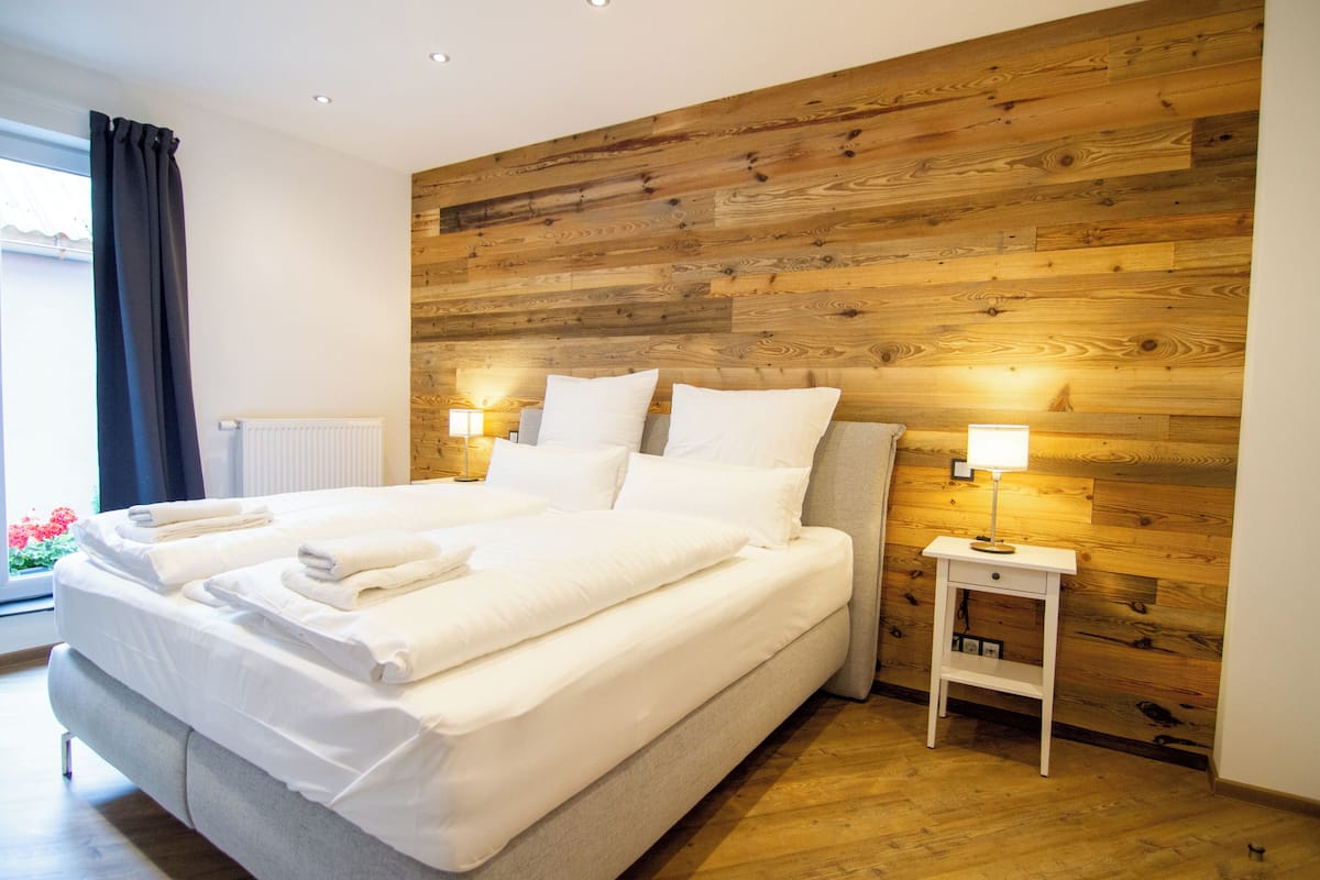 Een moderne hotelkamer met een groot bed, een wand van houten panelen, twee bedlampjes en uitzicht op bloemen door het raam.