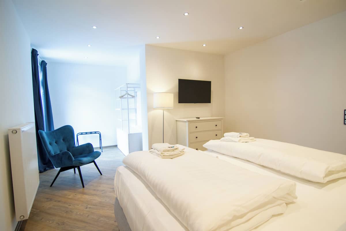 Een moderne hotelkamer met twee bedden, een blauwe fauteuil, een nachtkastje met een lamp en een flatscreen televisie aan de muur.