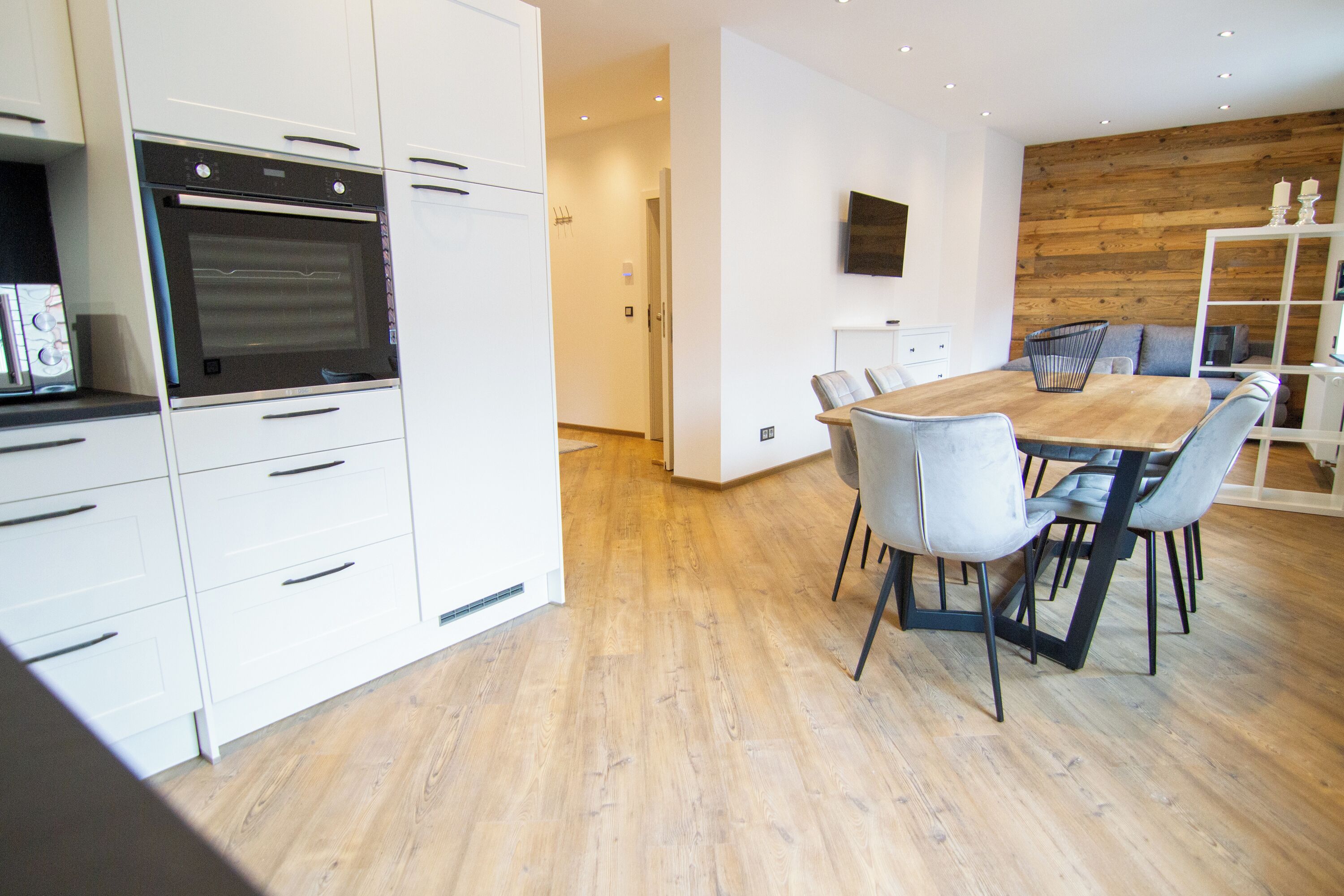 A modern kitchen with white cabinetry and a built-in oven. There is a dining area with a wooden table and chairs.