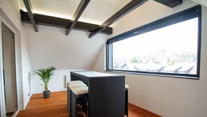 A small balcony with a wooden floor, a black wicker table with a cushion, a potted plant, and a large window offering a view of buildings. - Schiefer Suite Hotel & Apartment (Goslar)