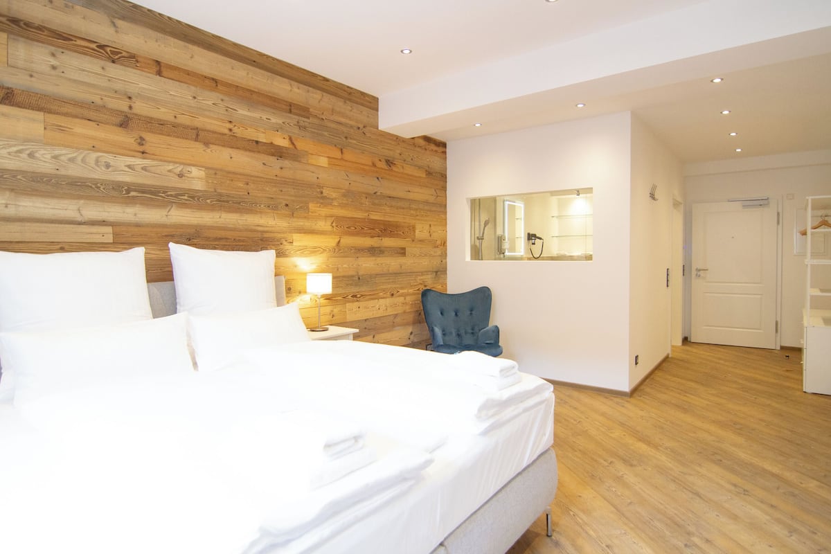 Een hotelkamer met een houten hoofdbord, een groot bed met witte lakens, een blauwe fauteuil, een spiegel en een kledingkast.