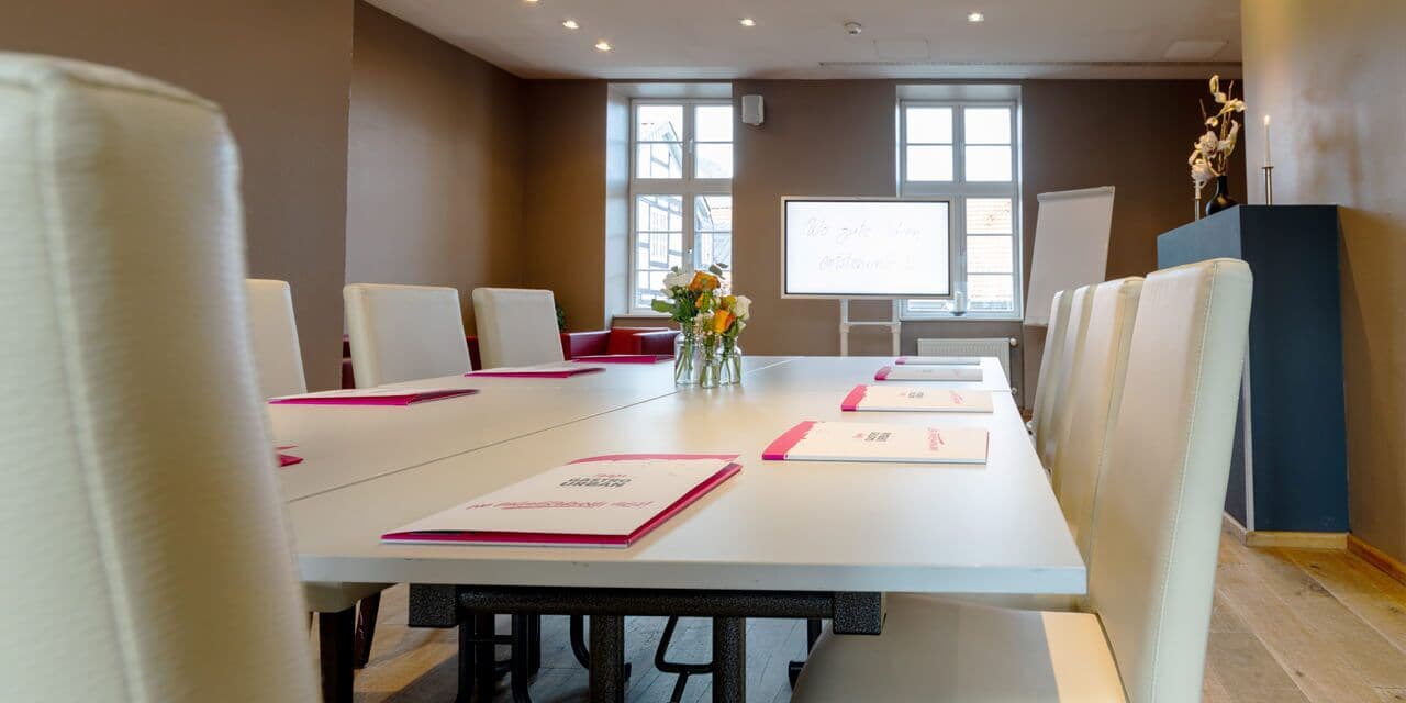Un salón de conferencias con una larga mesa, sillas blancas, una pizarra y un jarrón con flores.