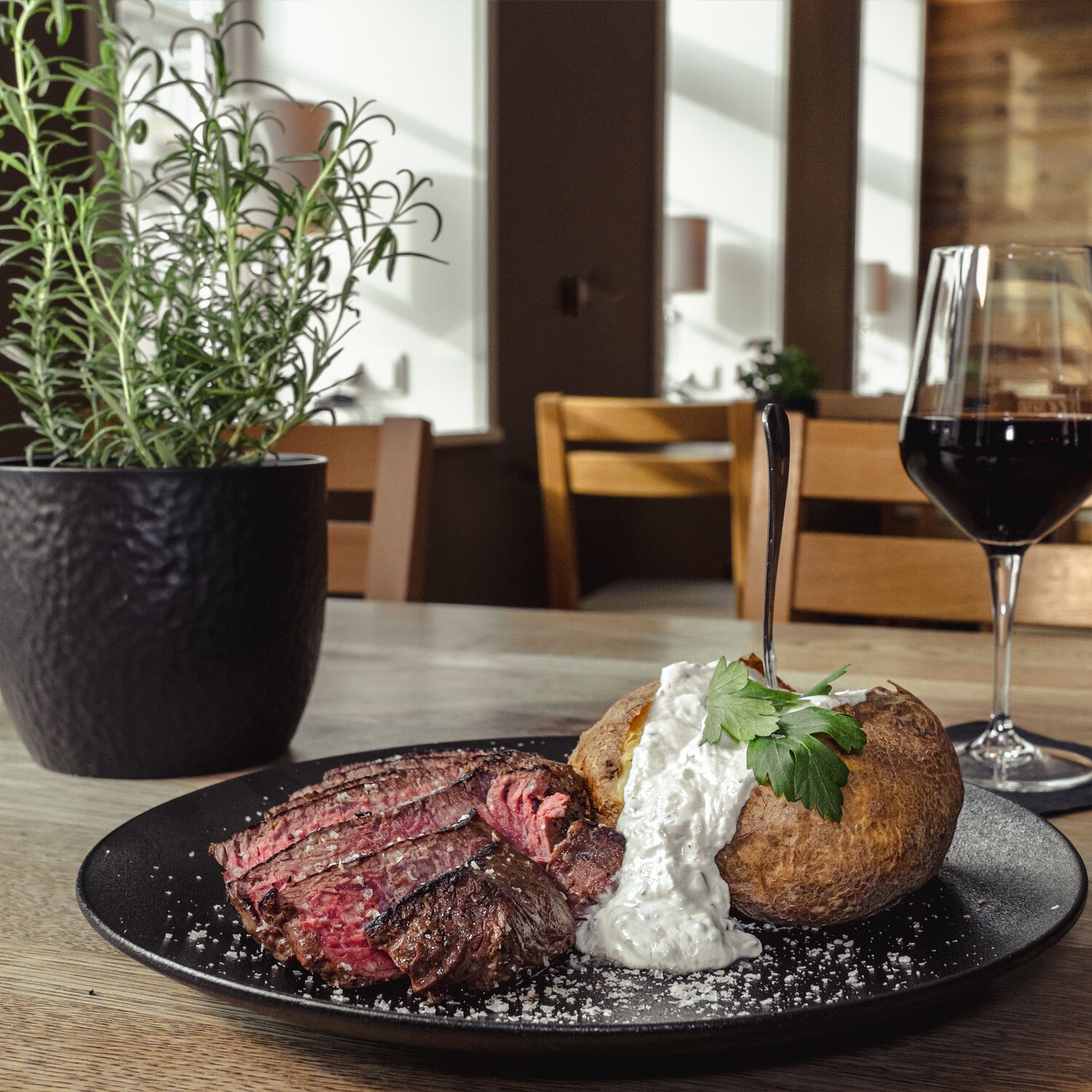 A plate with steak and a baked potato topped with sour cream and garnish, a glass of red wine, and a potted plant on a table.