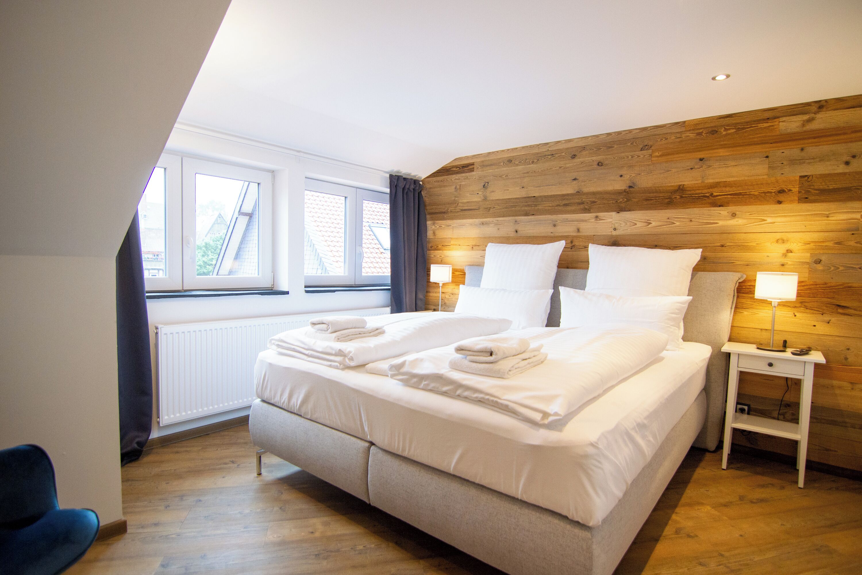 A modern bedroom with a large bed, wooden panel wall, two bedside lamps, and a view of the outside through a window.