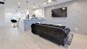 Una sala moderna con un sofá oscuro, un televisor montado en la pared y una cocina al fondo.