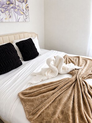 A neatly made bed with a beige headboard, white and black pillows, a beige blanket, and a white towel.