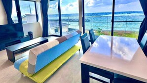 A modern living room with a sofa, dining table, and a TV. Large windows offer a view of the ocean. - AZ Hotel Ocean View (Amami)