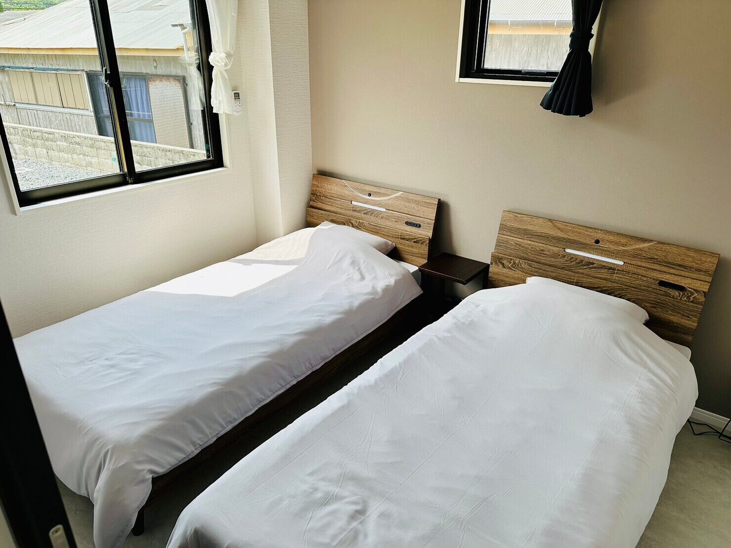 A small, clean room with two single beds, a wooden headboard, a nightstand, and a window with curtains.