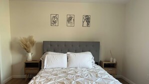 Ein Schlafzimmer mit einem gebürsteten Kopfteil, einer weißen Bettdecke, zwei Nachttischen mit Dekoration und drei gerahmten Kunstwerken an der Wand.