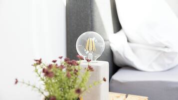 A clear glass light bulb on a white pedestal, a potted plant with purple flowers, and a grey sofa in the background.