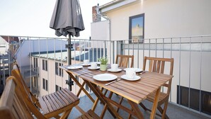 Mesa de madera dispuesta con platos, tazas y una planta pequeña en un balcón con baranda de metal y vistas a edificios.