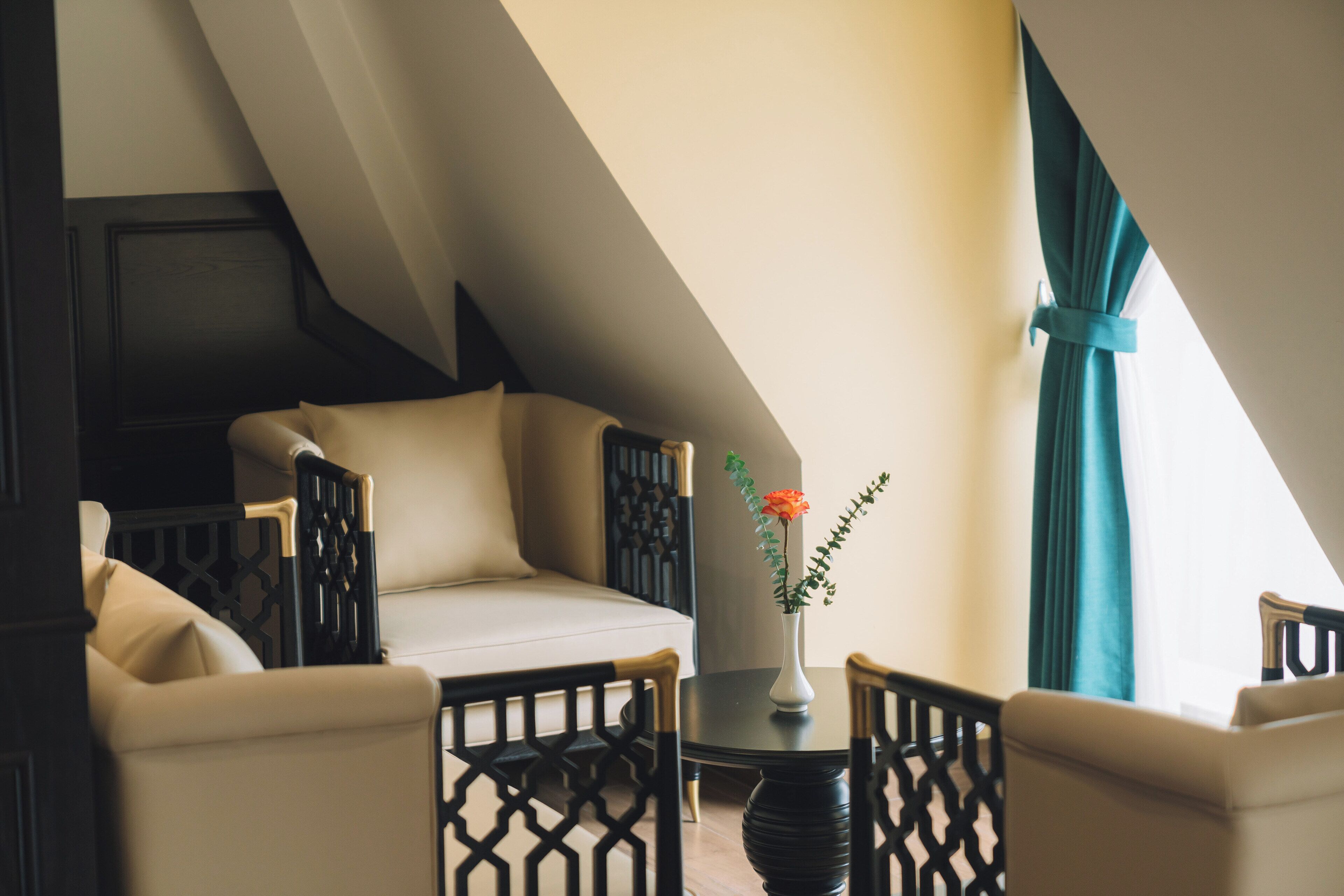 A cozy loft bedroom with a sloped ceiling, a sofa, two chairs, a small table with a vase of flowers, and a view of the sky through a window.