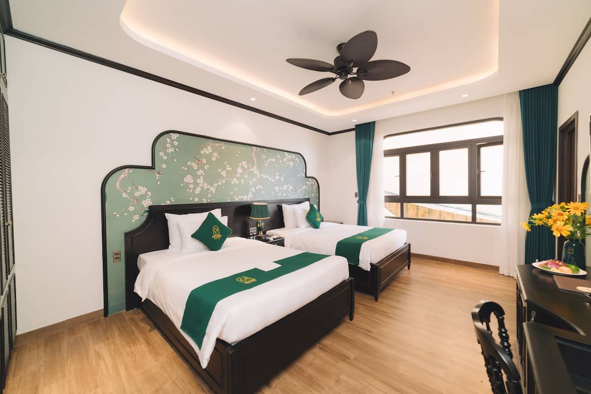 A hotel room with two beds, a ceiling fan, a large window, and a desk with a vase of flowers.