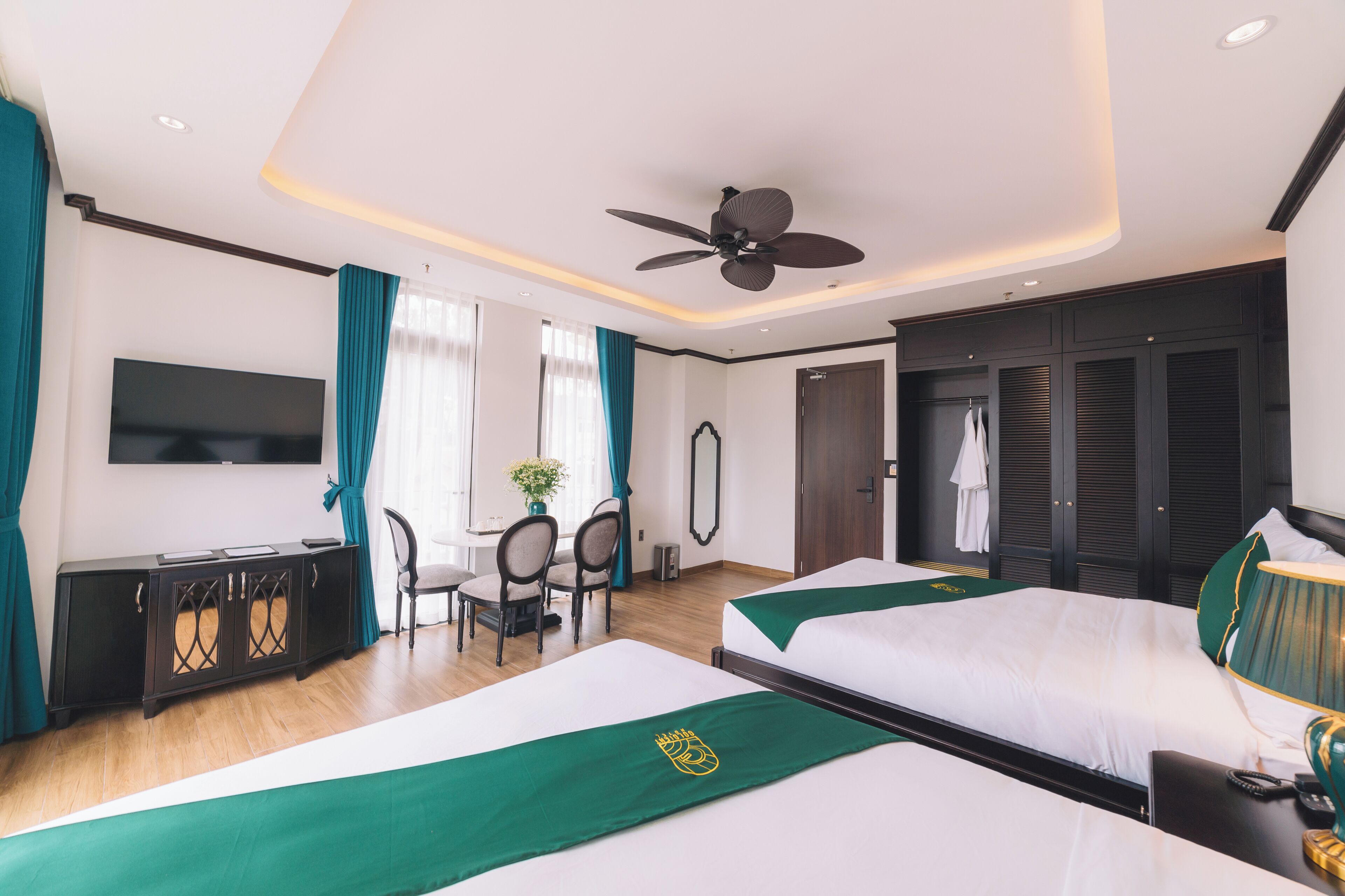 A hotel room with two beds, a desk, a chair, a TV, and a ceiling fan.