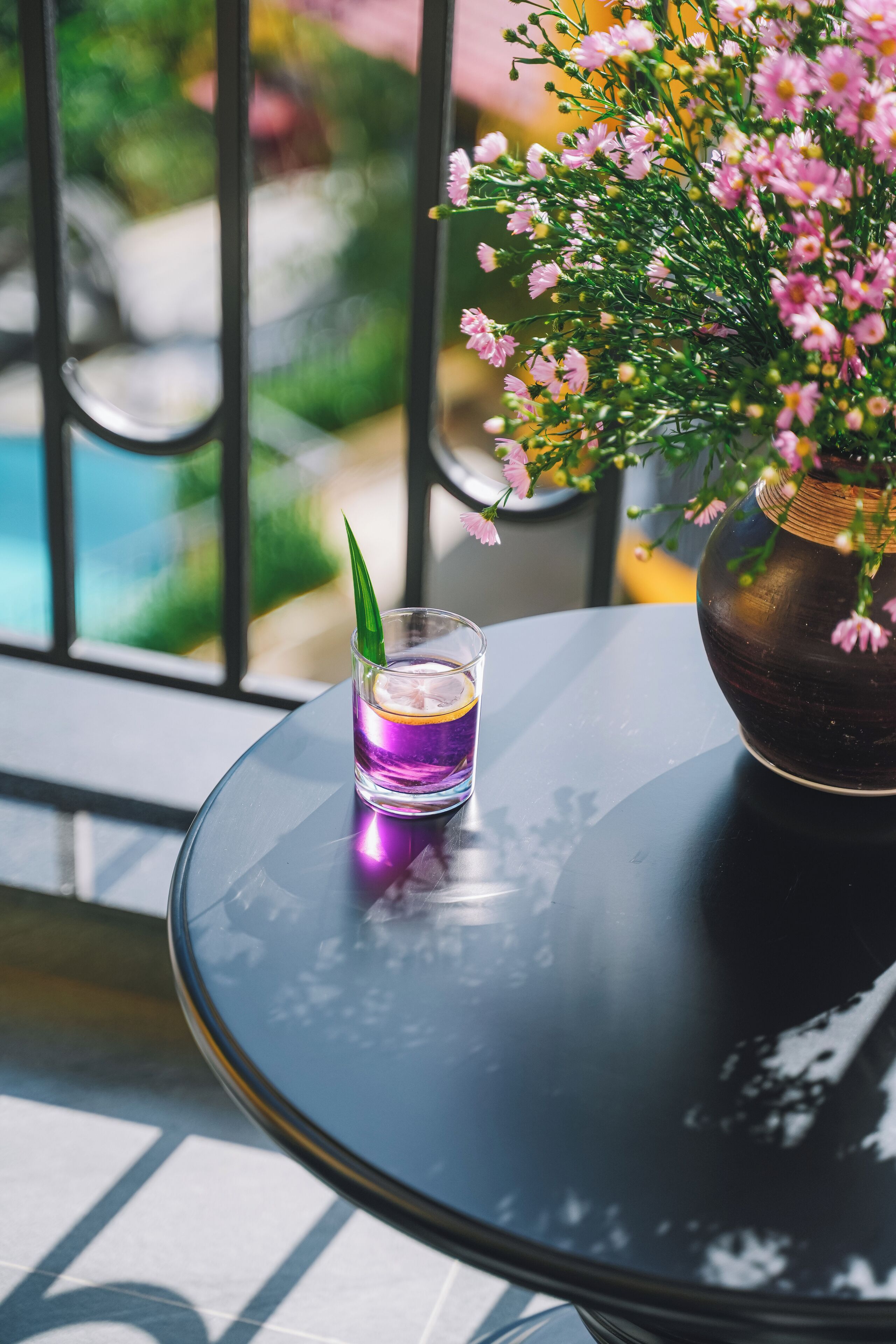 A glass of purple drink on a round table with a vase of pink flowers and a green leaf.