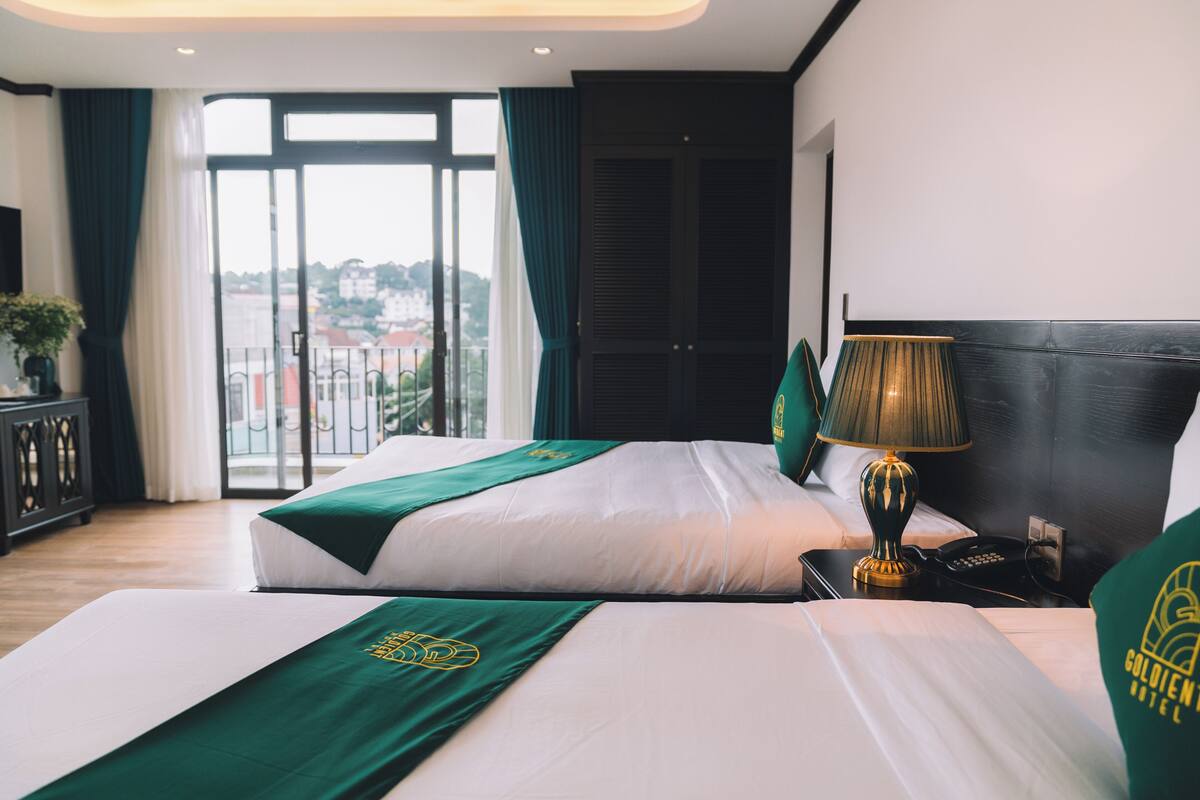 A hotel room with two beds, a large window with curtains, a nightstand with a lamp, and a view of buildings outside.