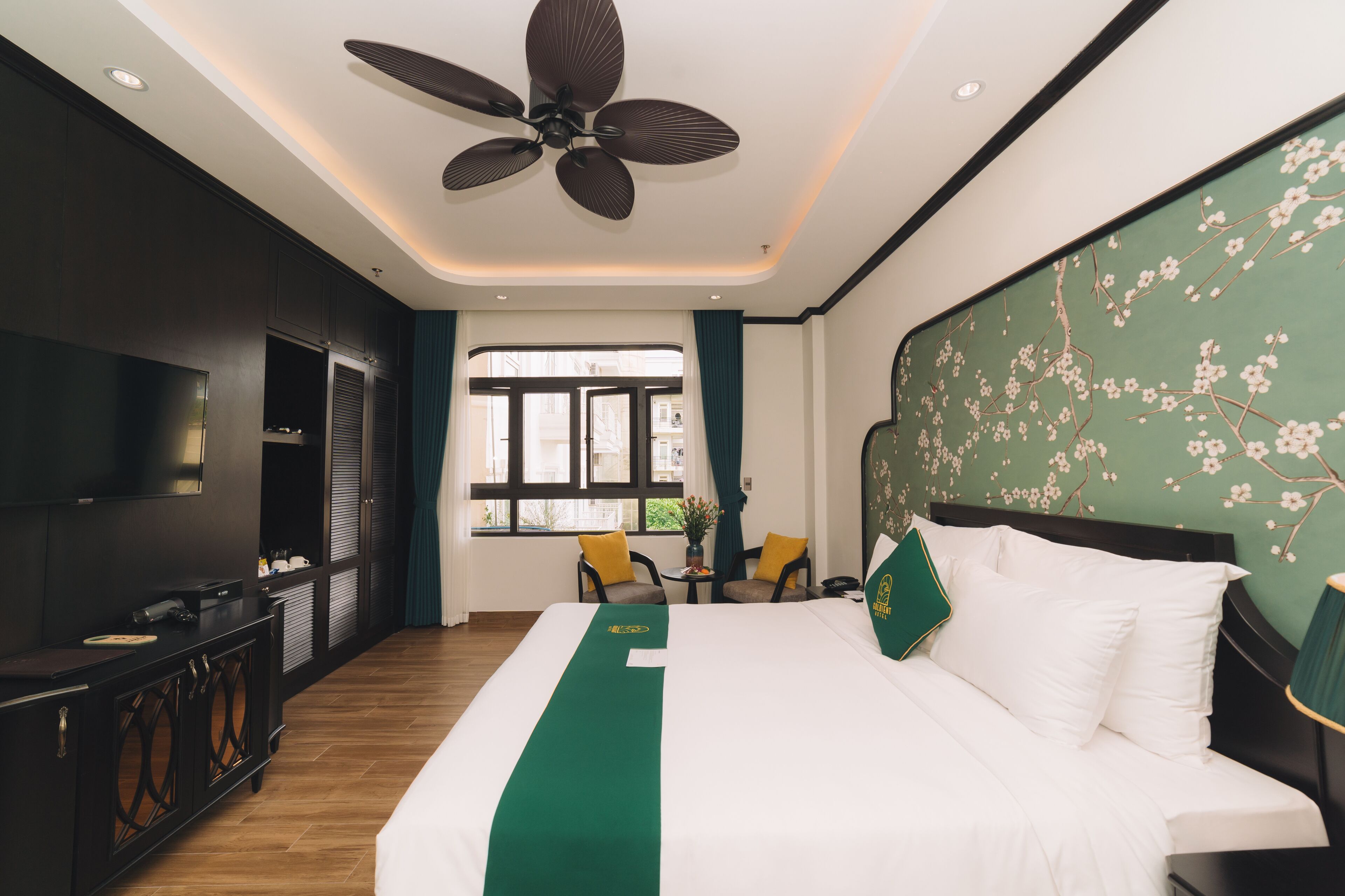 A hotel room with a large bed, a ceiling fan, a TV, a desk with a computer, and a window with a view of the outside.
