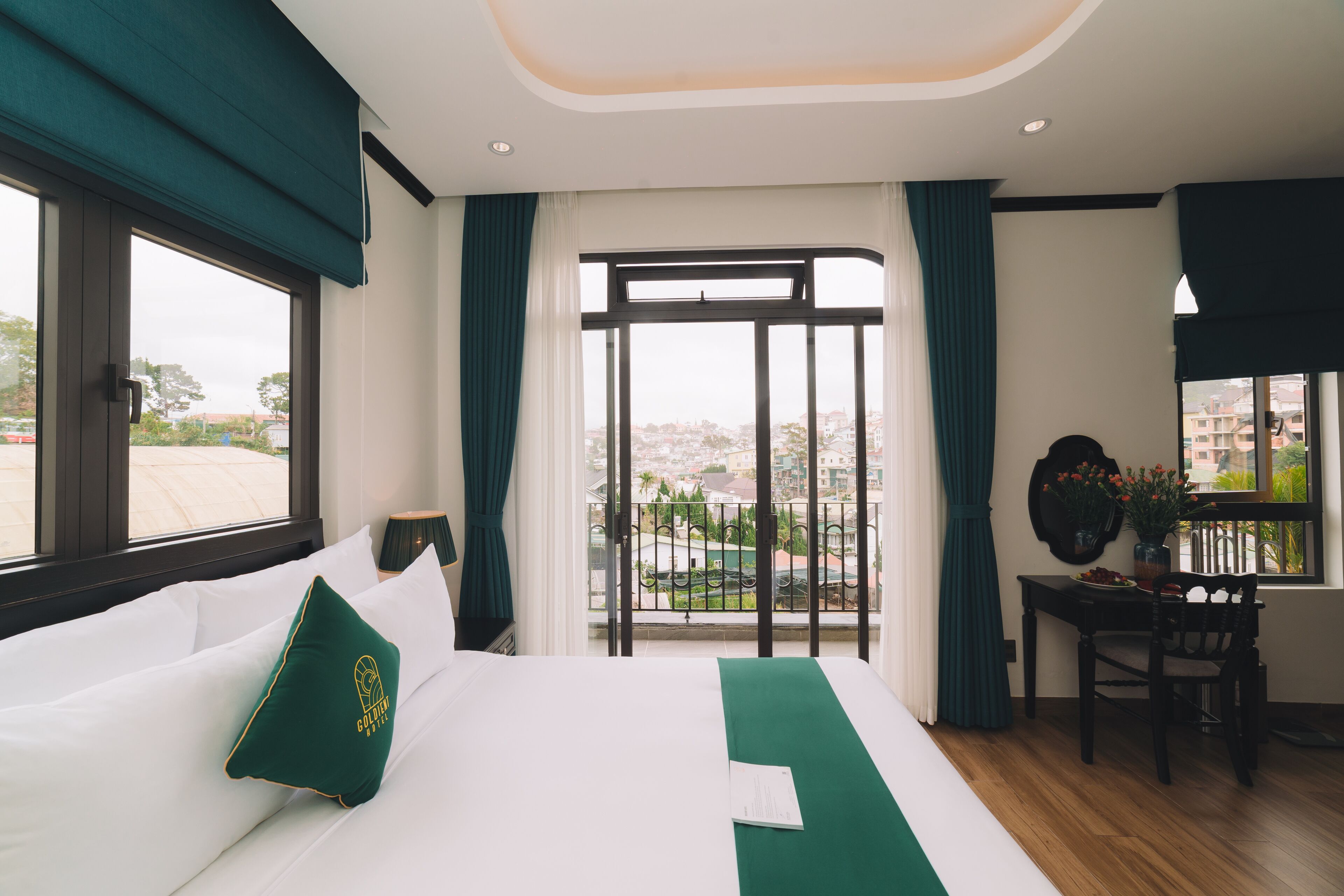 A hotel room with a large bed, a view of the city through a balcony, and a small table with a vase of flowers.