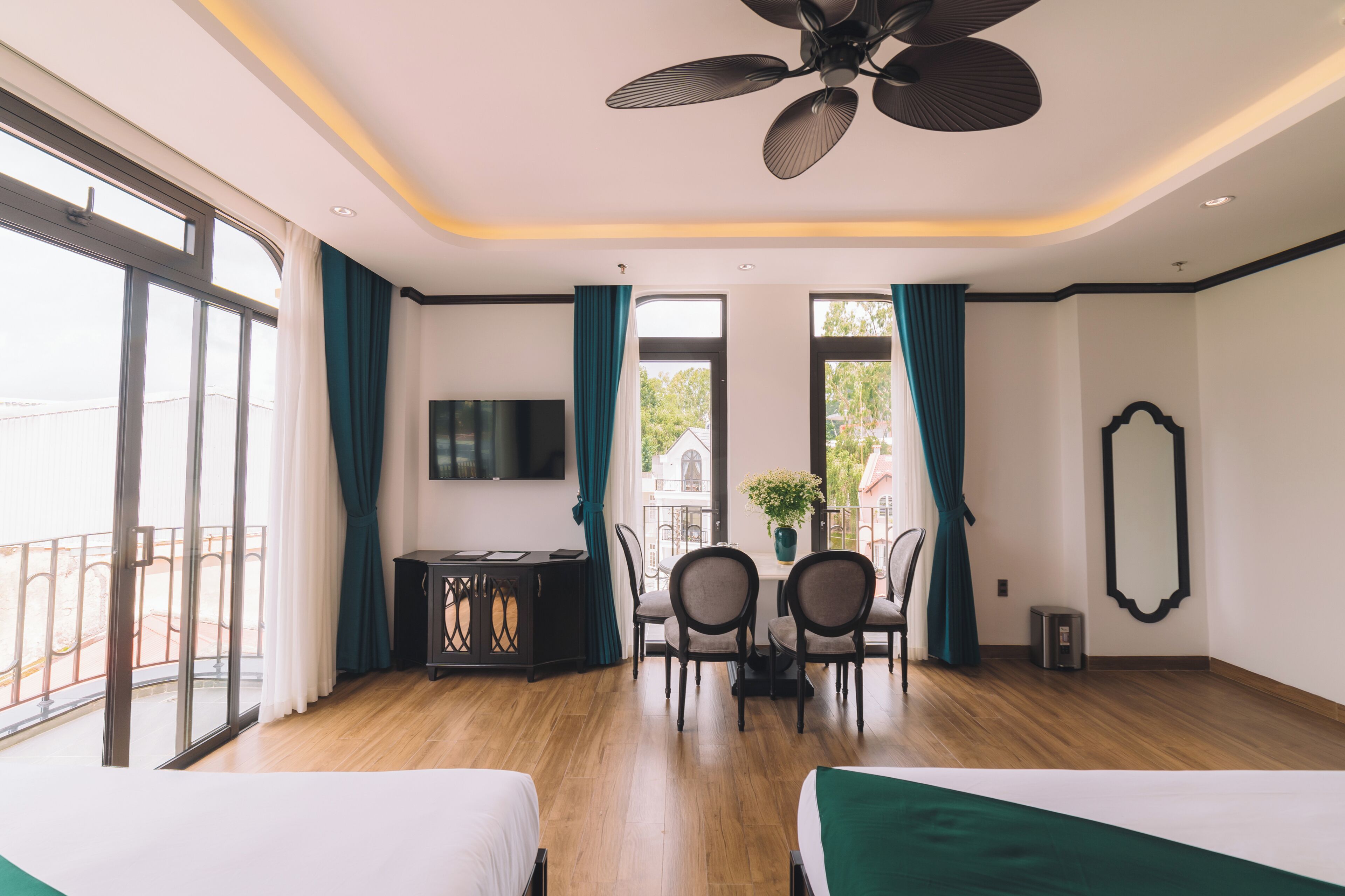 A hotel room with a large bed, a smaller bed, a dining table with chairs, a TV, and a balcony with a railing.