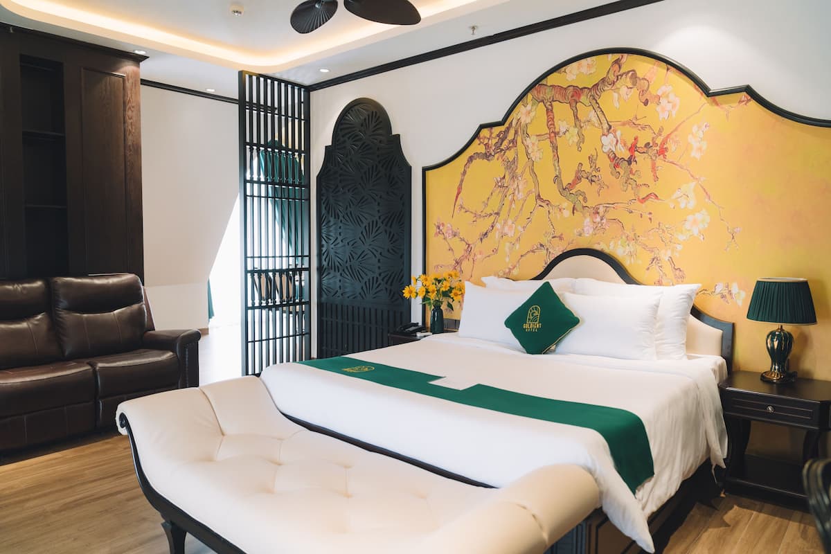 A hotel room with a large bed, a sofa, a decorative wall, and a nightstand with a lamp.
