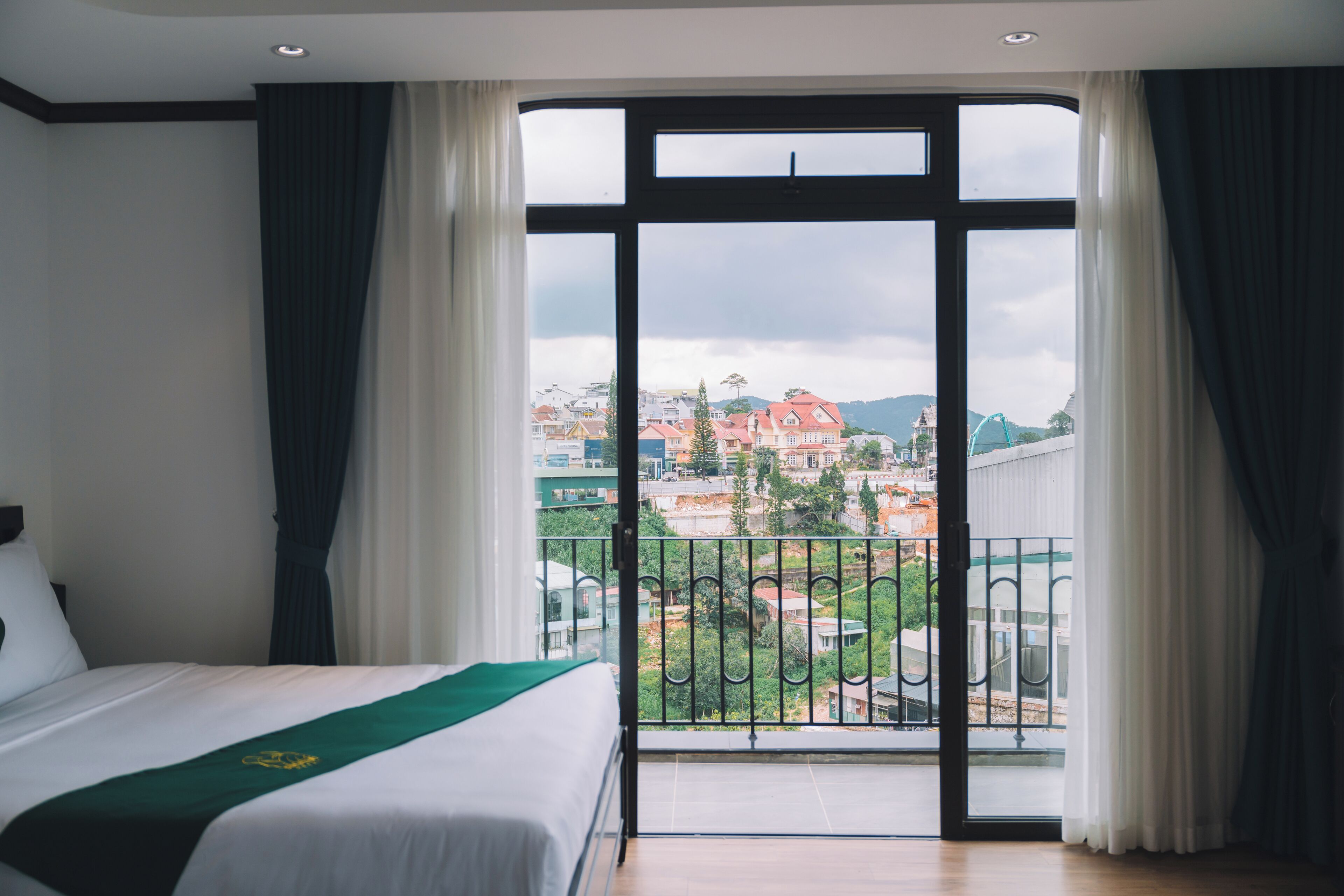 A hotel room with a large window, a bed with a green bedspread, and a view of a cityscape.