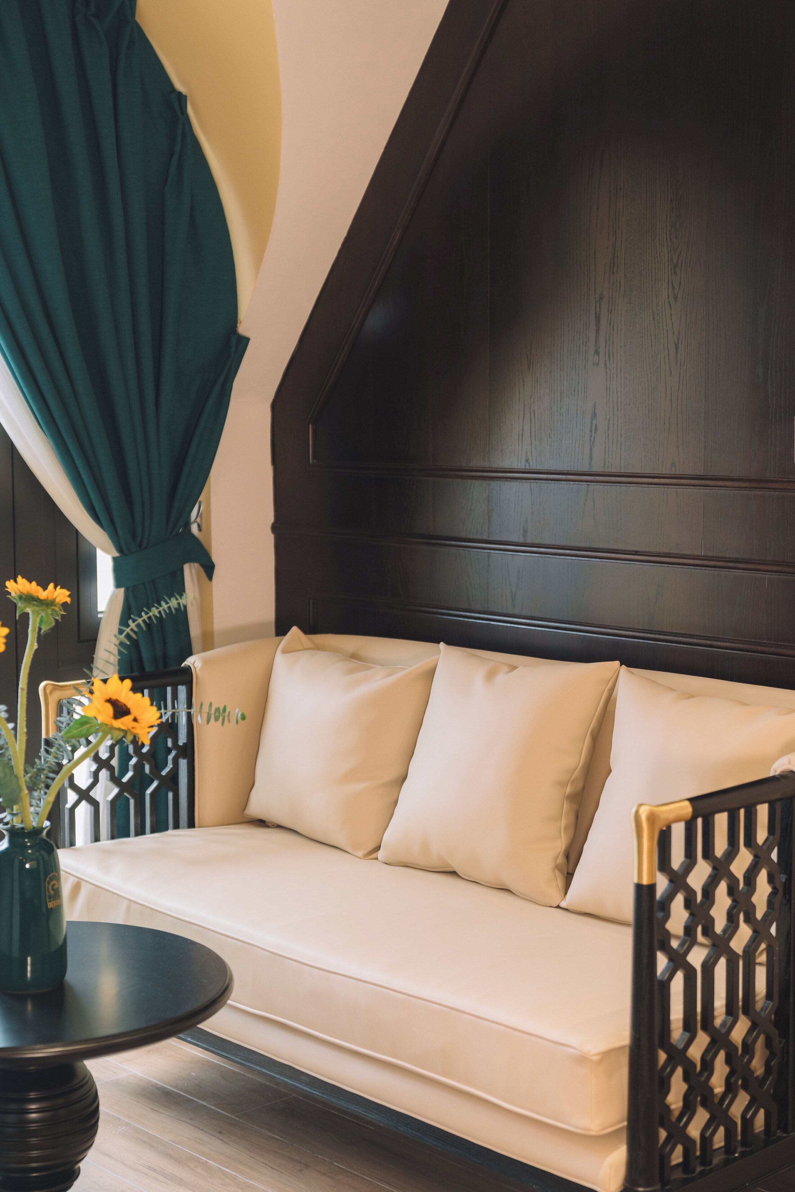 A sofa with two pillows, a small round table with a vase of flowers, and a dark wooden headboard.