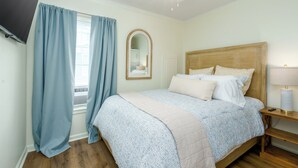 A bedroom with a bed, a nightstand, a lamp, a ceiling fan, a TV, and curtains. - Cozy cottage with easy access to beach & restaurants- Pet Friendly! (PASS CHRISTIN)
