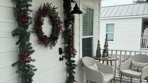 A porch with a white door, a wreath, and Christmas decorations. There are wicker chairs and a small table with a 'Merry Christmas' pillow. - Cozy cottage with easy access to beach & restaurants- Pet Friendly! (PASS CHRISTIN)
