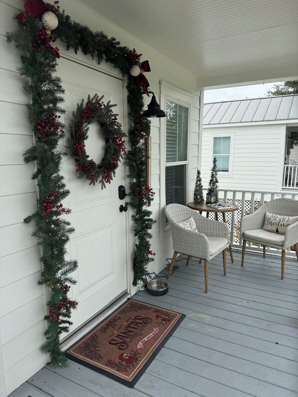 A porch with a white door, a wreath, and Christmas decorations. There are wicker chairs and a small table with a 'Merry Christmas' pillow.