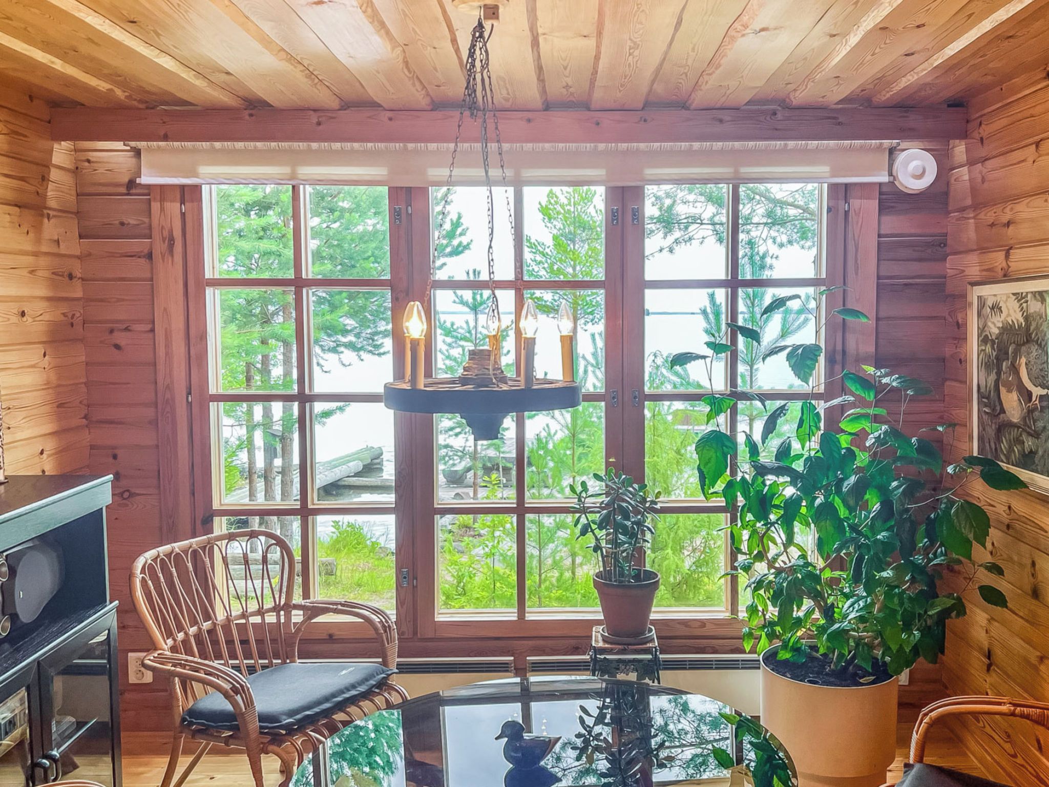 Un accogliente interno di una baita con pareti in legno, un'ampia finestra vista su un paesaggio verde, una poltrona in vimini, un tavolo con un oggetto decorativo e una lampada a sospensione.