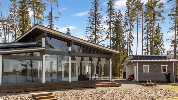 Et moderne hus med store glasvinduer, en træterrasse og en stikvej af sten, der fører til indgangen.