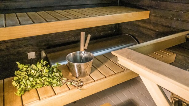 Een sauna met houten banken, een metalen wasbak en een houten plank met een bos groene bladeren.