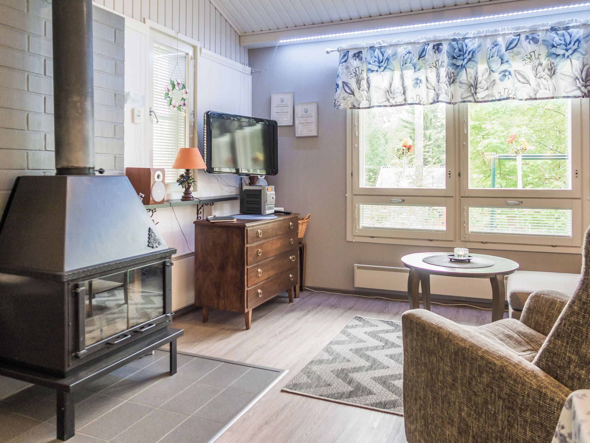 Un salon chaleureux avec une cheminée, un téléviseur, une commode en bois et une fenêtre donnant sur un paysage verdoyant.