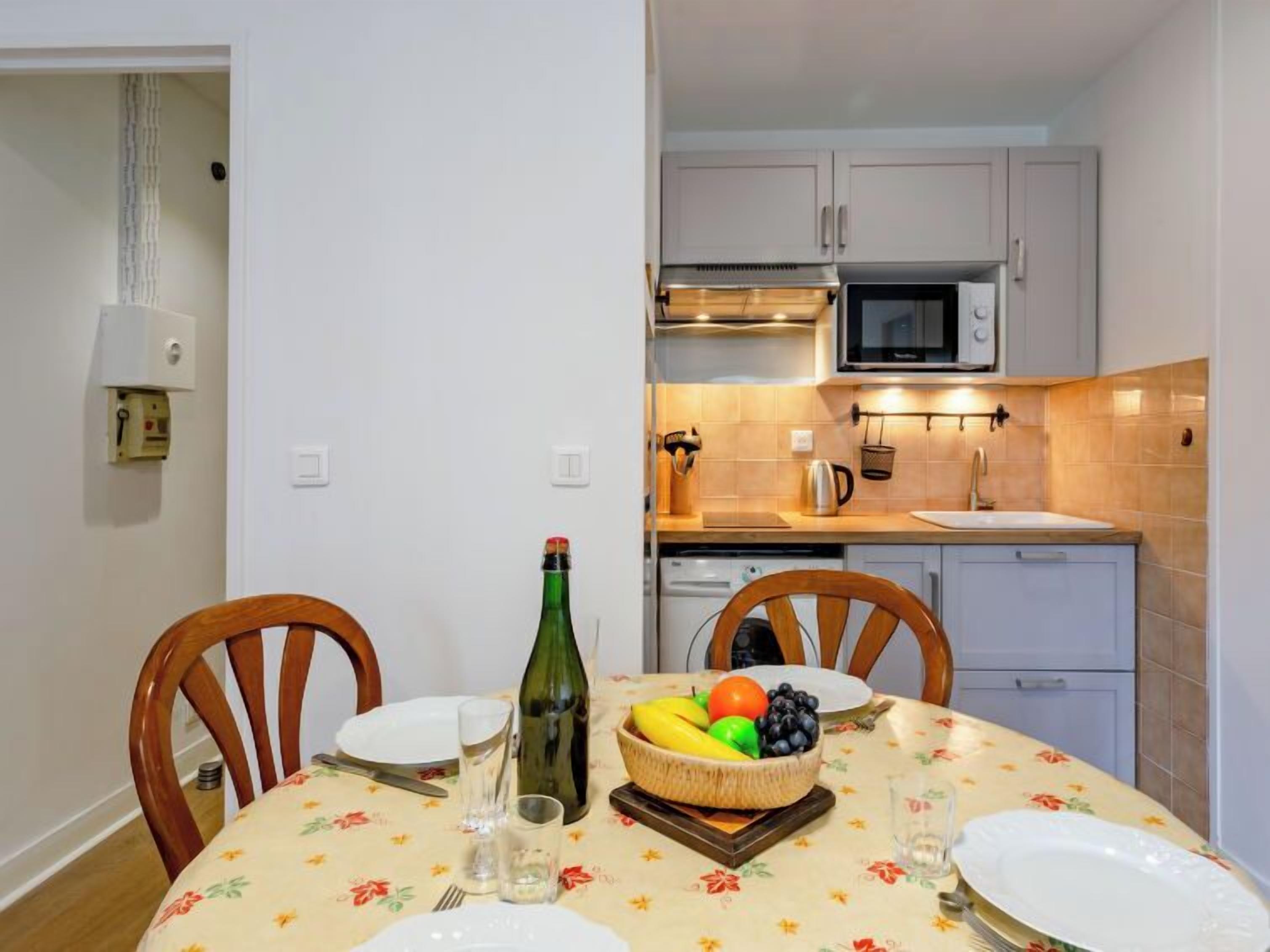 A dining area with a table set for two, a bottle of wine, and a fruit bowl.