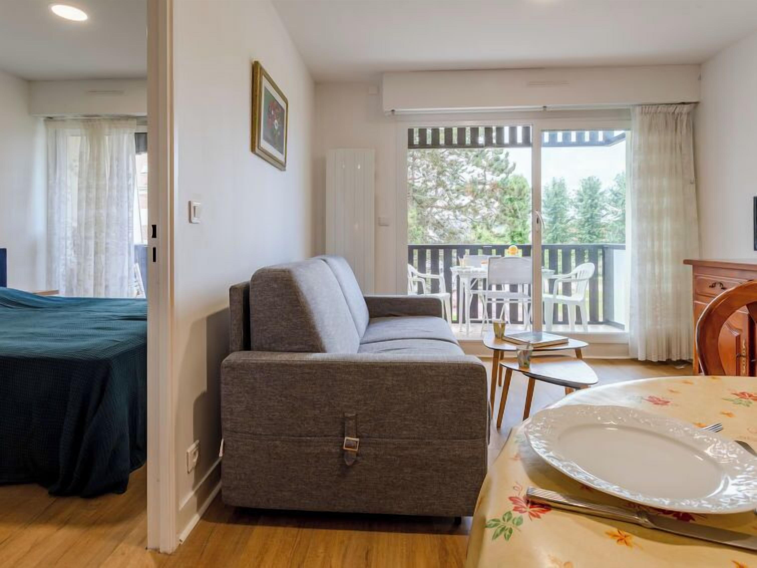 Ein Zimmer mit einer Couch, einem Esstisch mit Teller und Besteck, einem kleinen Tisch mit Stühlen und Blick durch eine Schiebetür auf einen Außenbereich mit Tisch und Stühlen.