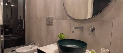 A modern bathroom with a round mirror, a green bowl sink, a bidet, and a towel rack.