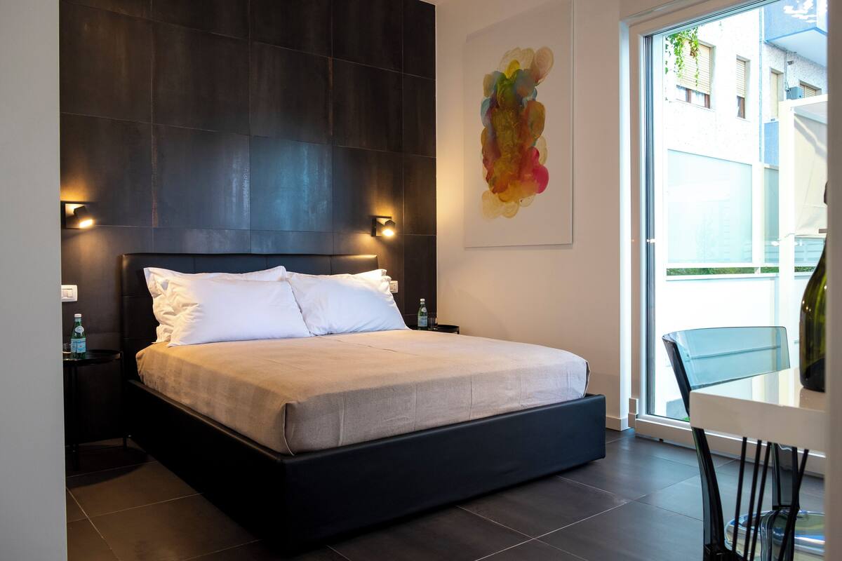 Un dormitorio moderno con una cama grande, paneles de pared oscuros, una mesita de vidrio y un cuadro abstracto colorido.