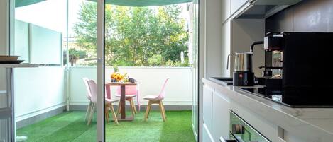Una cocina moderna con comedor, un portón corredizo de vidrio que da a un balcón con césped artificial, y vistas a árboles y edificios.