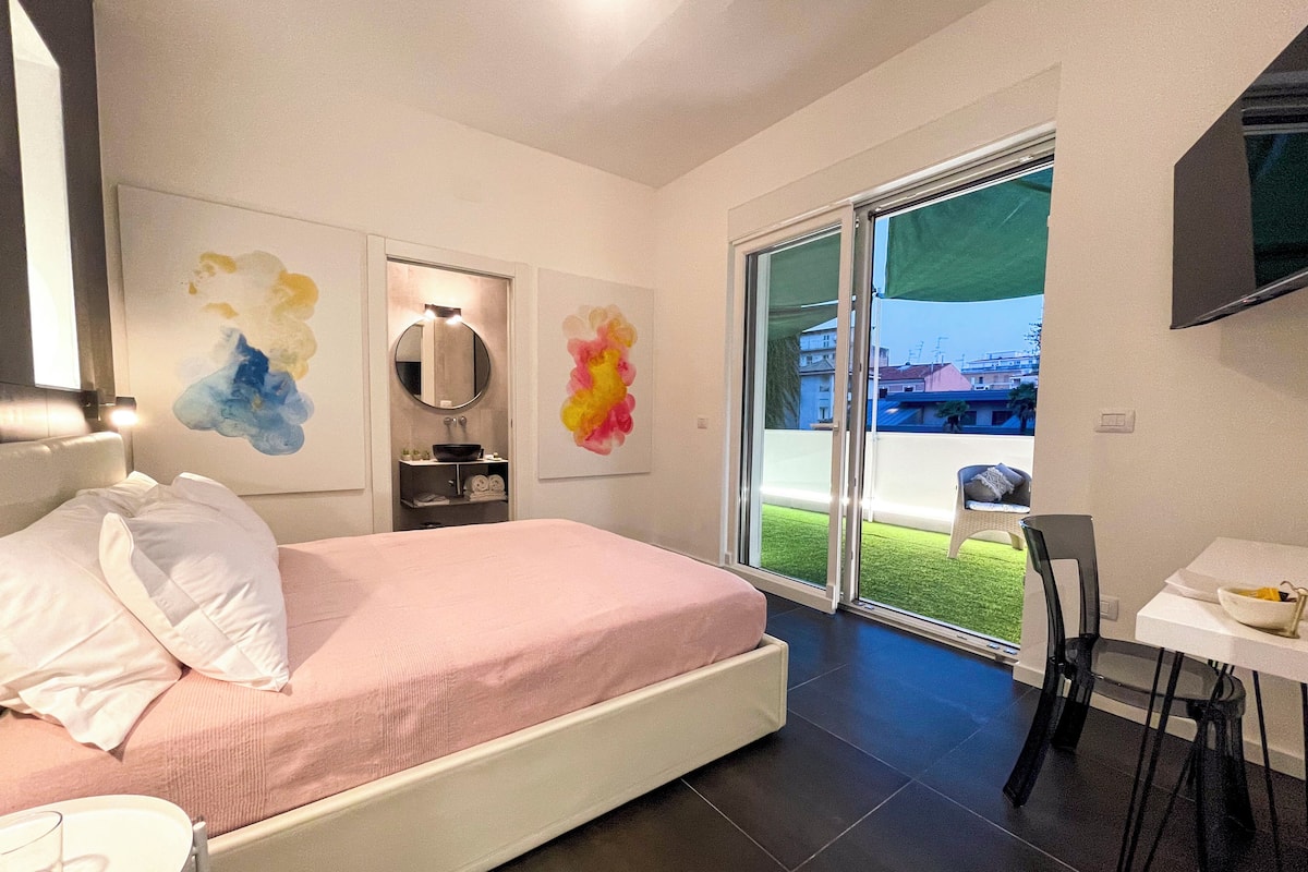 Un dormitorio moderno con cama, una mesita, una silla, un baño con espejo y un balcón con vistas a edificios.