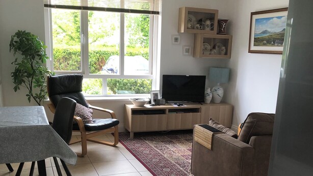 Ein Wohnzimmer mit Fernseher, einer Couch, einem Esstisch und einem großen Fenster mit Jalousien.