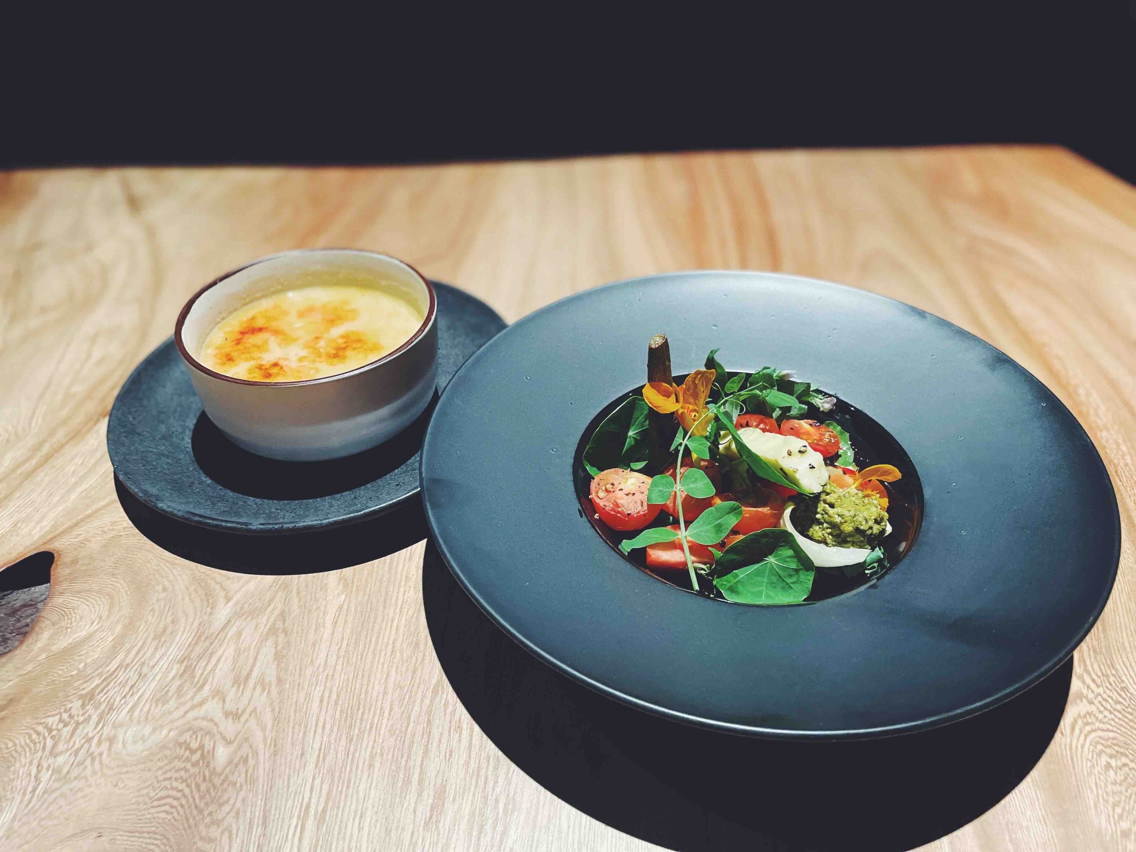 A dark blue plate with a salad and a small bowl of soup on a wooden table.