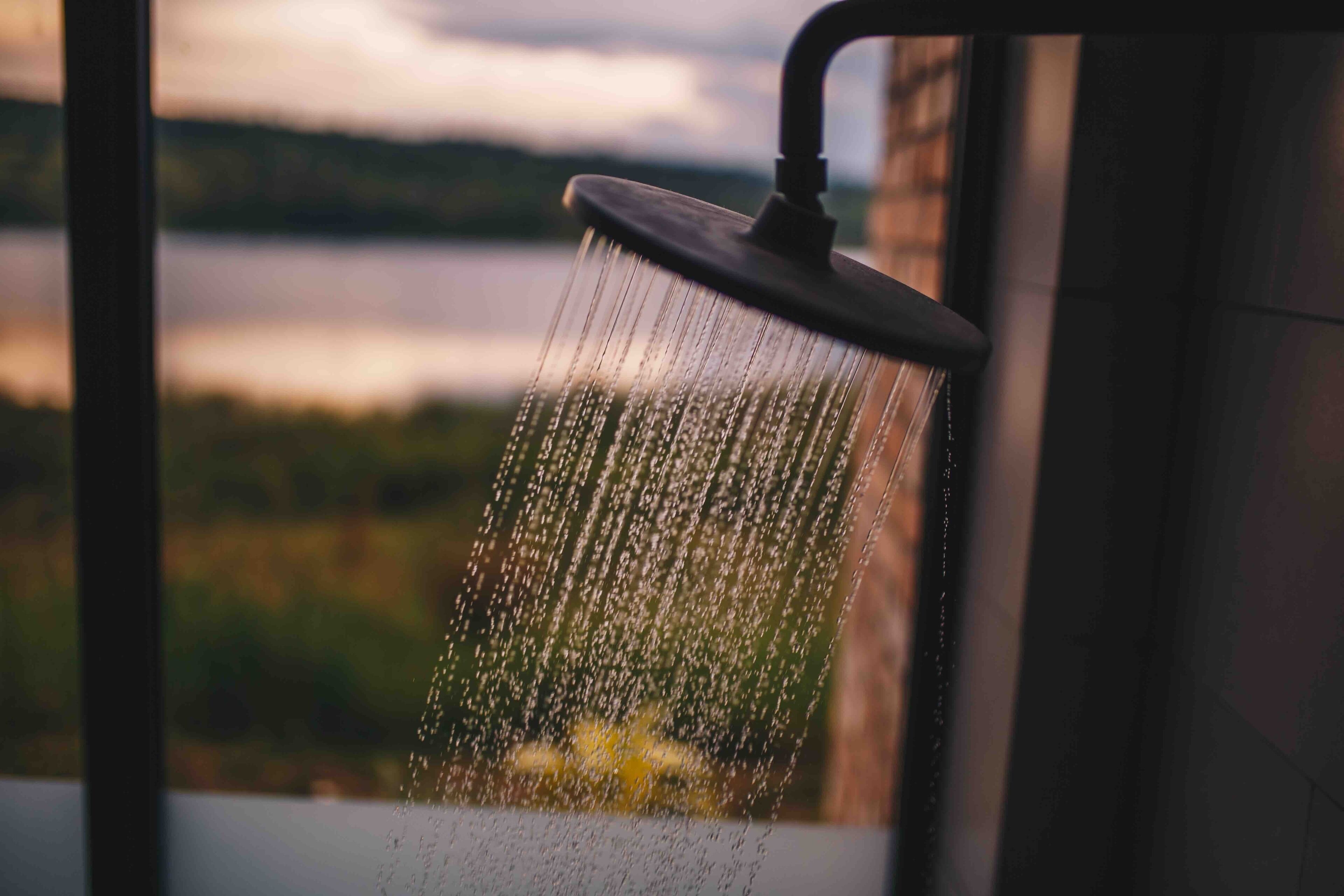 Un pommeau de douche laissant couler l’eau, vu à travers une fenêtre donnant sur un plan d’eau et une végétation luxuriante.