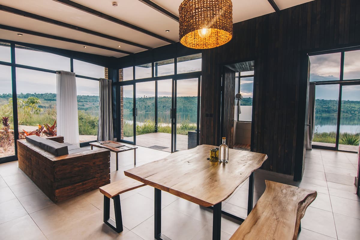 A spacious room with a large wooden table, a bench, and a sofa. There are large windows offering a view of a lake and greenery.